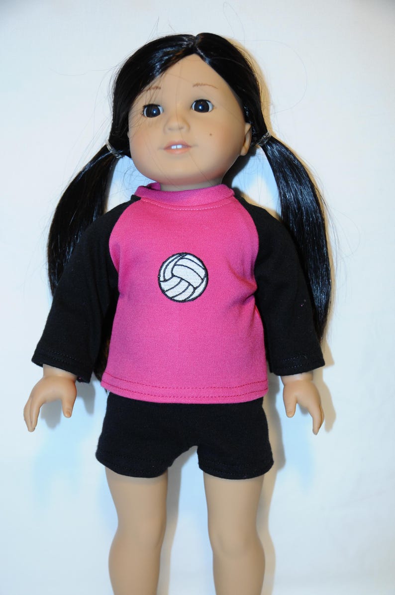 Puede incluir: Una mu&ntilde;eca con cabello largo y negro peinado en dos coletas, que viste una camiseta rosa de manga larga con mangas negras y un gr&aacute;fico de voleibol blanco en el pecho, junto con pantalones cortos negros. La mu&ntilde;eca tiene piel clara y ojos oscuros.