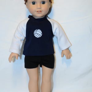 Puede incluir: Una mu&ntilde;eca con cabello casta&ntilde;o rojizo recogido en un mo&ntilde;o, que viste una camiseta de manga larga azul marino y blanca con un gr&aacute;fico de voleibol blanco en el pecho, combinada con pantalones cortos negros lisos. La mu&ntilde;eca tiene piel clara y ojos azules.