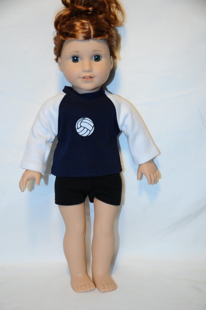 Puede incluir: Una mu&ntilde;eca con cabello casta&ntilde;o rojizo recogido en un mo&ntilde;o, que viste una camiseta de manga larga azul marino y blanca con un gr&aacute;fico de voleibol blanco en el pecho, combinada con pantalones cortos negros lisos. La mu&ntilde;eca tiene piel clara y ojos azules.