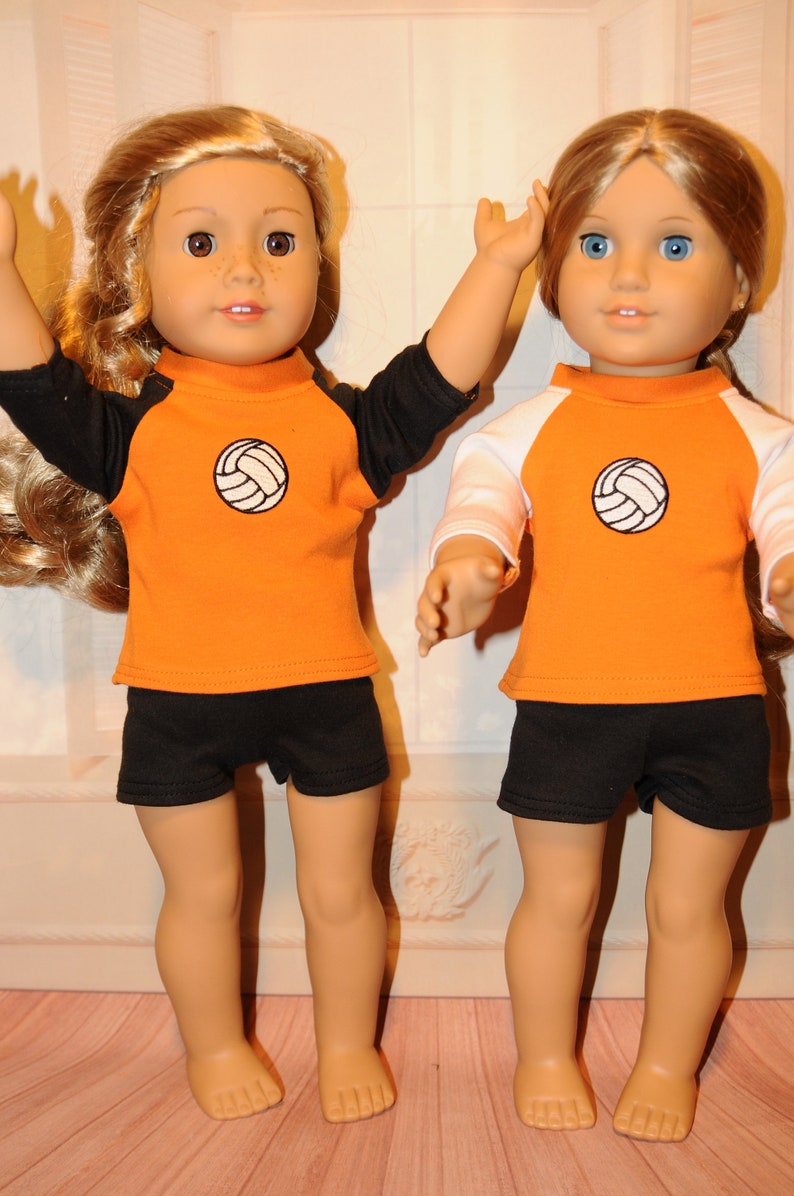 Puede incluir: Dos mu&ntilde;ecas de piel clara y cabello largo con ropa deportiva. La mu&ntilde;eca de la izquierda lleva una camiseta naranja con mangas negras y pantalones cortos negros. La mu&ntilde;eca de la derecha lleva una camiseta naranja con mangas blancas y pantalones cortos negros. Ambas camisetas tienen un gr&aacute;fico de voleibol.