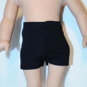 Puede incluir: Unos pantalones cortos negros lisos usados por una mu&ntilde;eca. Los pantalones cortos presentan un dise&ntilde;o sencillo y ajustado con una cintura alta y aberturas de pierna rectas. La tela parece ser un material mate y liso que cubre el torso inferior.