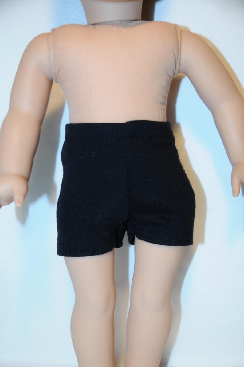Puede incluir: Unos pantalones cortos negros lisos usados por una mu&ntilde;eca. Los pantalones cortos presentan un dise&ntilde;o sencillo y ajustado con una cintura alta y aberturas de pierna rectas. La tela parece ser un material mate y liso que cubre el torso inferior.
