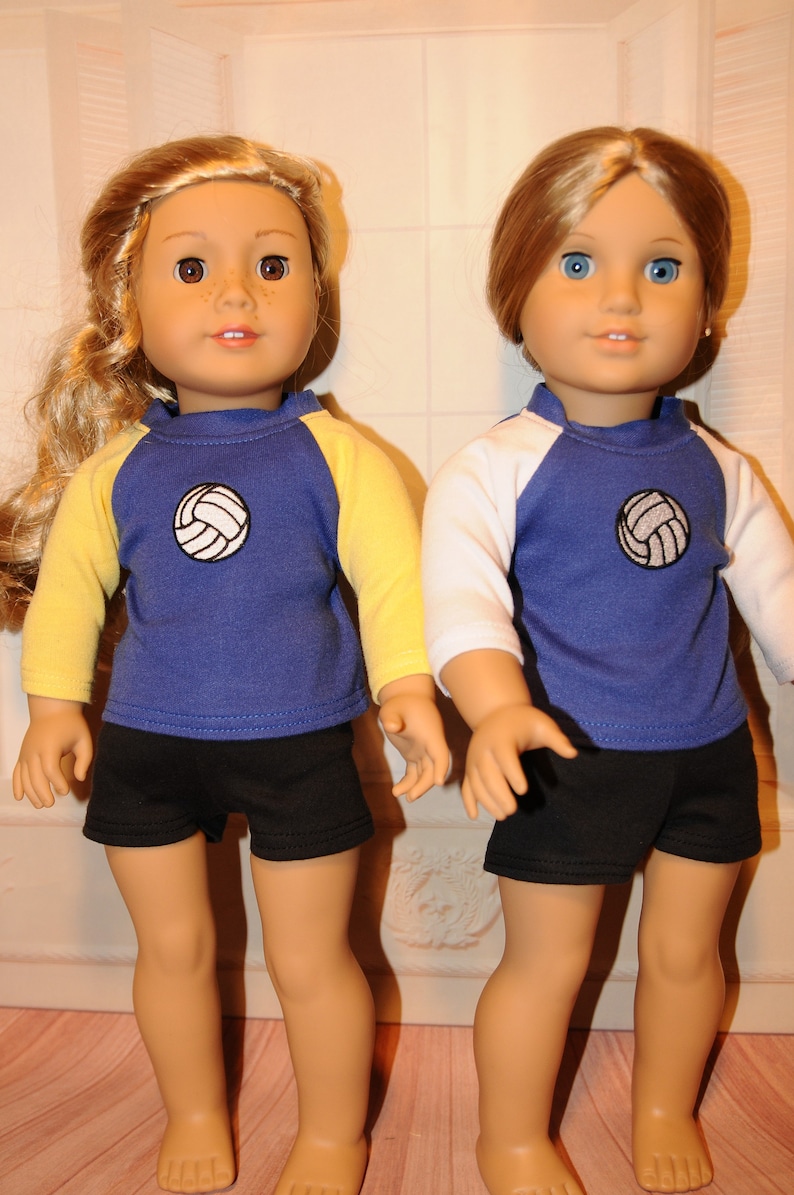 Puede incluir: Dos mu&ntilde;ecas de piel clara con ropa deportiva. La mu&ntilde;eca de la izquierda tiene el pelo largo y rubio y lleva una camiseta azul con mangas amarillas y pantalones cortos negros. La mu&ntilde;eca de la derecha tiene el pelo casta&ntilde;o y lleva una camiseta azul con mangas blancas y pantalones cortos negros. Ambas camisetas tienen un gr&aacute;fico de voleibol blanco en el pecho.