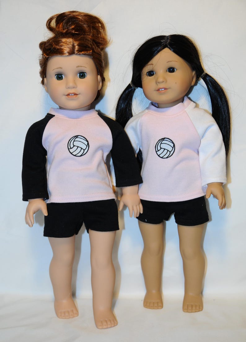 Puede incluir: Dos mu&ntilde;ecas de pie una al lado de la otra, ambas con camisetas de color rosa claro con un gr&aacute;fico de voleibol negro en el pecho. La mu&ntilde;eca de la izquierda tiene el pelo casta&ntilde;o rojizo y mangas negras, mientras que la de la derecha tiene el pelo negro y mangas blancas. Ambas mu&ntilde;ecas llevan pantalones cortos negros.