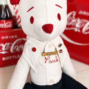 May include: A white plush bear with red ears, red eyes, and a red nose. The bear wears a white collared shirt with the name Paul embroidered in red thread, a gold tie bar, three small pins, and solid black pants. The bear has a soft fabric texture.