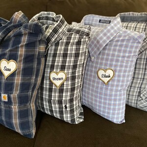 May include: Several plaid shirt pillows in various colors, including blue, black, and gray. Each pillow has a heart-shaped patch with a name embroidered on it. The pillows are arranged on a brown surface.