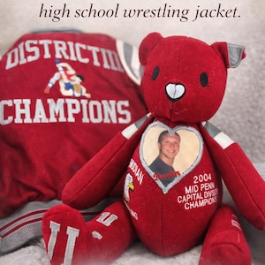 May include: A red memory bear made from a high school wrestling jacket. The bear has a heart-shaped patch with a photo and text that reads "2004 MID PENN CAPITAL DIVISION CHAMPIONS". The jacket is red with white and gray accents.