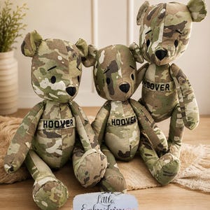 May include: Three handmade teddy bears in a camouflage pattern. Each bear has a black heart-shaped nose and the name "HOOVER" embroidered on a patch. The bears are sitting on a light brown surface, with a small sign that reads "Little Embroiderers".