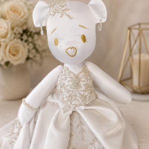 May include: A white satin plush bear wearing a formal gown with intricate silver embroidery and beadwork. The bear features gold embroidered eyes and a heart-shaped nose, a beaded bracelet, and decorative silver hair and ear embellishments.