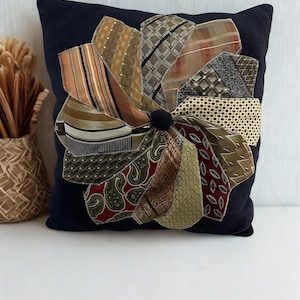 May include: A square navy blue throw pillow featuring a central circular applique made from various neckties arranged in a sunburst pattern. The neckties display diverse patterns including stripes, paisley, polka dots, and geometric shapes in earth tones.
