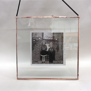 Puede incluir: Un marco de fotos de vidrio de tono cobre con un cord&oacute;n de colgar negro. El marco contiene una fotograf&iacute;a en blanco y negro de dos ni&ntilde;os de pie frente a una chimenea de ladrillo. Los ni&ntilde;os llevan atuendos a juego y sonr&iacute;en.