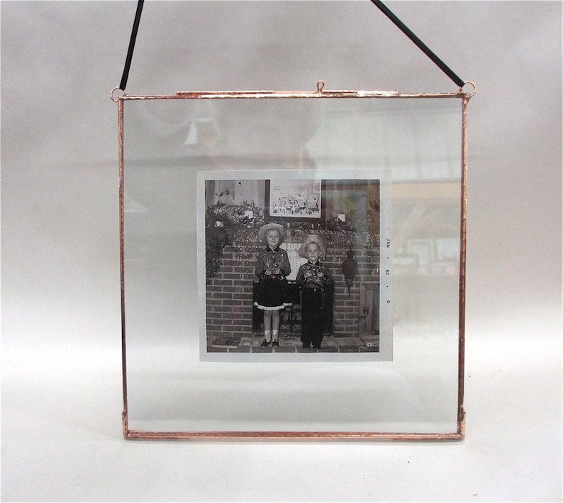 Puede incluir: Un marco de fotos de vidrio de tono cobre con un cord&oacute;n de colgar negro. El marco contiene una fotograf&iacute;a en blanco y negro de dos ni&ntilde;os de pie frente a una chimenea de ladrillo. Los ni&ntilde;os llevan atuendos a juego y sonr&iacute;en.