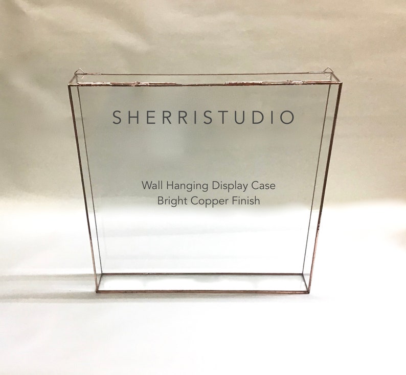 May include: A clear glass wall hanging display case with a bright copper finish. The case has a shelf and is labeled "SHERRISTUDIO Wall Hanging Display Case Bright Copper Finish".