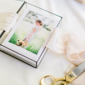 May include: A glass box with a black frame containing a photo album with a photo of a person in a white dress holding a bouquet of flowers. A piece of white fabric and a pair of gold scissors are on the table.