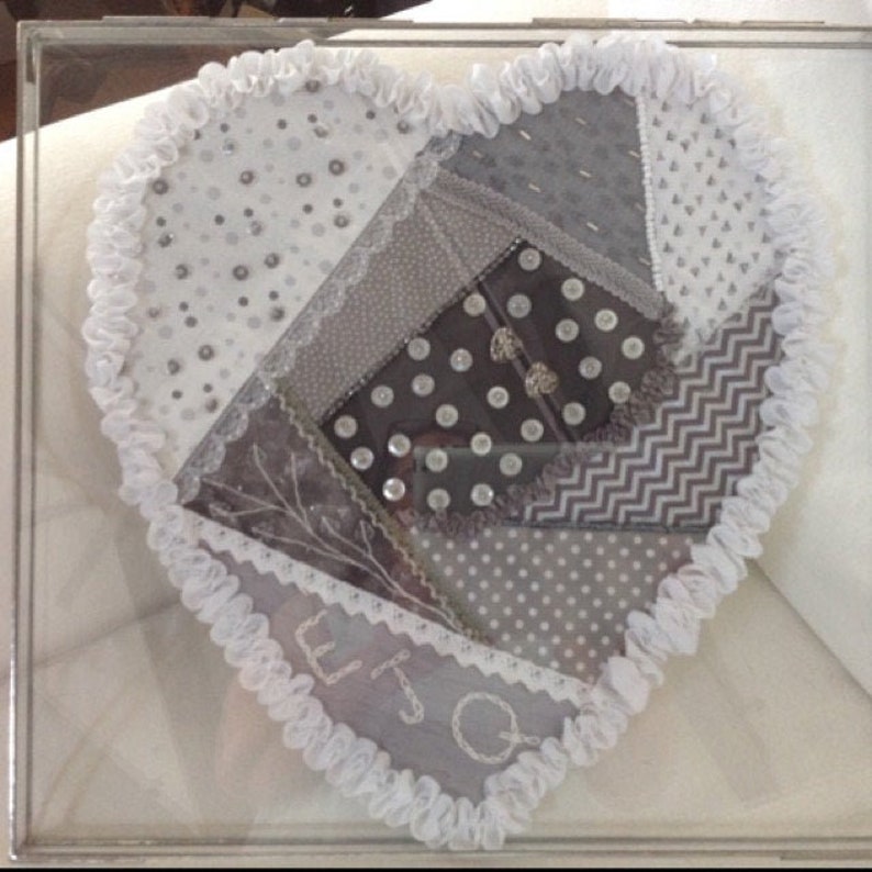 May include: A gray and white heart-shaped quilt with a white lace border. The quilt is made of different fabrics with various patterns, including polka dots, stripes, and zigzags. The word "EtQ" is embroidered in the lower right corner.