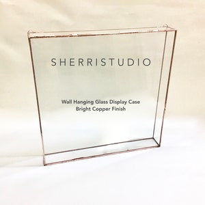 May include: A square wall-mounted glass display case with a bright copper finish. The case has a clear glass front and sides with a copper frame. The text "SHERRISTUDIO" and "Wall Hanging Glass Display Case Bright Copper Finish" is printed on the glass.