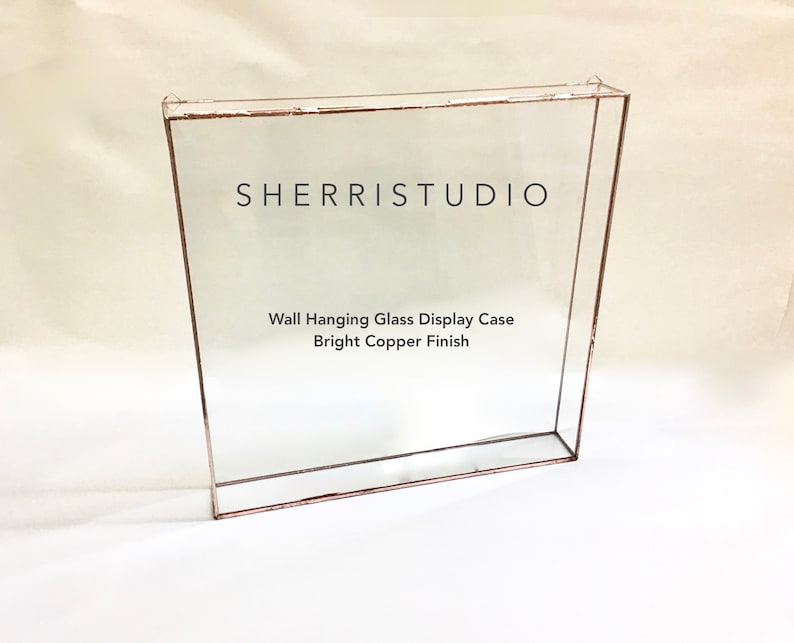 May include: A square wall-mounted glass display case with a bright copper finish. The case has a clear glass front and sides with a copper frame. The text "SHERRISTUDIO" and "Wall Hanging Glass Display Case Bright Copper Finish" is printed on the glass.