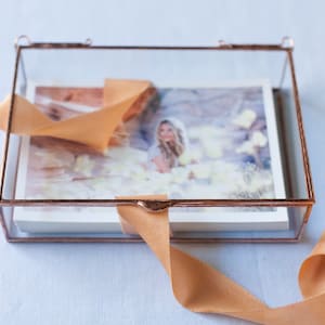 May include: A glass box with a copper frame holds a printed photograph of a woman in a white dress. The box is decorated with a light brown ribbon.