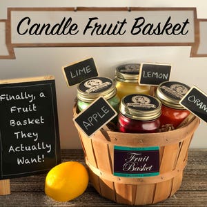 May include: A wooden basket filled with candles shaped like fruit. The candles are labeled "Lime", "Apple", "Lemon", and "Orange". A chalkboard sign reads "Finally, a Fruit Basket They Actually Want!"