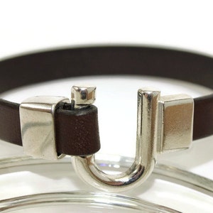 May include: A brown leather bracelet with a silver clasp. The clasp is a U-shaped design with a small silver bar on the top.