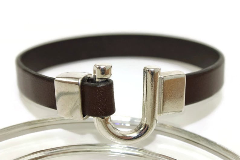 May include: A brown leather bracelet with a silver clasp. The clasp is a U-shaped design with a small silver bar on the top.
