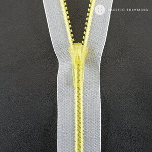 May include: A close-up of a yellow zipper with a white tape. The zipper is closed and the slider is in the center. The zipper is on a black background.