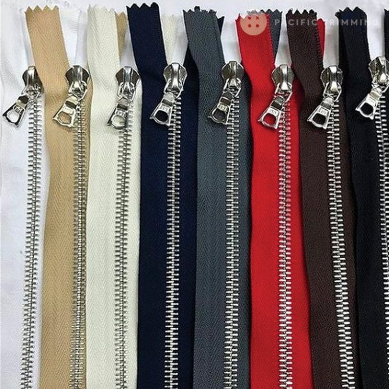K&ouml;nnte beinhalten: Eine Auswahl an farbigen Rei&szlig;verschl&uuml;ssen in verschiedenen Farbt&ouml;nen, darunter Wei&szlig;, Beige, Marineblau, Grau, Rot, Braun und Schwarz. Jeder Rei&szlig;verschluss hat einen silbernen Metallschieber und Z&auml;hne. Die Rei&szlig;verschl&uuml;sse sind vertikal angeordnet.