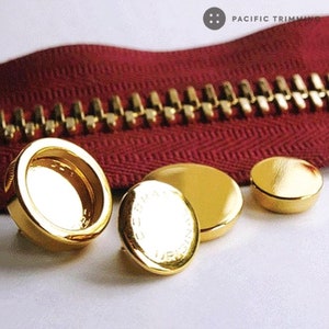 May include: Three gold-coloured metal zipper pulls. The largest pull is round with a raised edge and a smooth surface. The middle pull is round with a raised edge and a textured surface with lettering. The smallest pull is round and smooth.