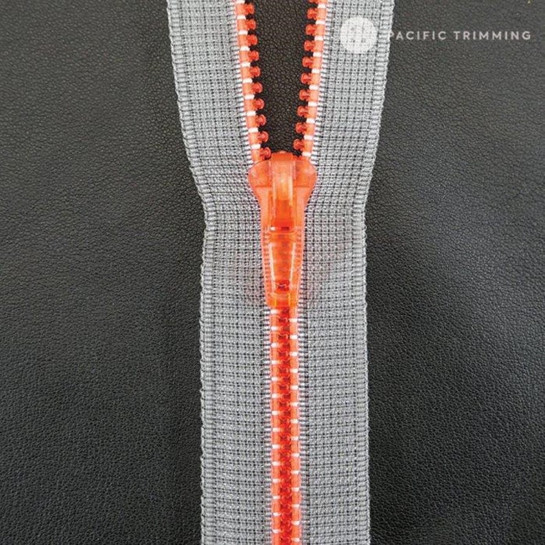 May include: A close-up of a zipper with a gray tape and an orange slider. The zipper is open and the slider is in the middle of the tape.