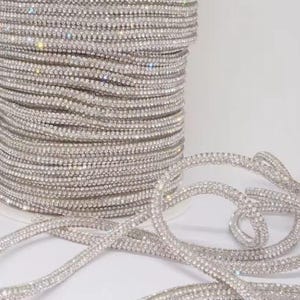 May include: A spool of silver-toned rhinestone trim featuring multiple rows of small, reflective crystals set in a flexible band. The trim is coiled tightly on a cylindrical spool, with several loose strands extending outward across the surface.