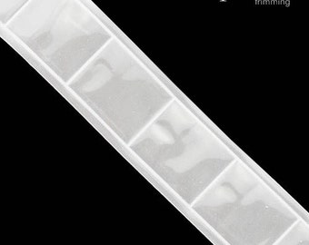 Könnte beinhalten: Weißer Besatz mit einem sich wiederholenden Muster aus durchsichtigen, quadratischen Verzierungen. Der Besatz ist aus einem durchsichtigen Stoff.