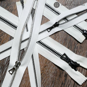 Op de afbeelding: Witte ritsband met zwarte tanden en zilveren schuifjes. De ritsband ligt op een houten oppervlak.