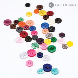 Può includere: Assortimento di bottoni di plastica colorati con quattro fori ciascuno. I bottoni sono in varie tonalità di rosso, blu, verde, giallo, viola, rosa, marrone e bianco. I bottoni sono disposti su una superficie bianca. PACIFIC TRIMMING è stampato nell'angolo in alto a destra dell'immagine.