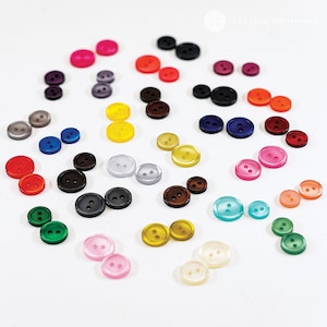 May include: Assortment of colorful plastic buttons in various shades, including red, blue, green, yellow, and brown. The buttons are arranged on a white surface.