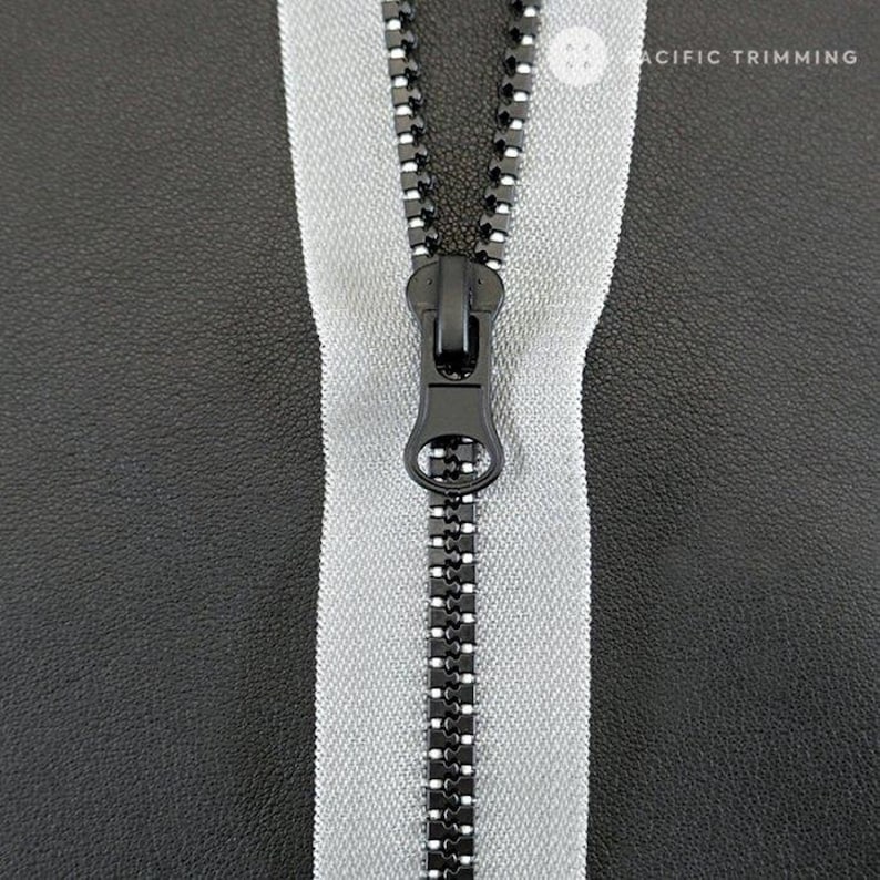 May include: A close-up of a silver zipper with a black zipper pull. The zipper is on a black fabric background.