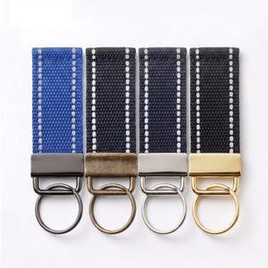 May include: Four rectangular fabric keychains with reflective white stitching. The first is blue with gunmetal hardware, the second is black with antique brass hardware, the third is black with silver hardware, and the fourth is black with gold hardware.