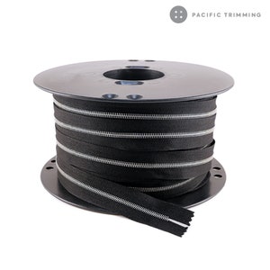 May include: A spool of black zipper tape with silver teeth. The tape is wound around a black plastic spool. The spool is labeled "PACIFIC TRIMMING".