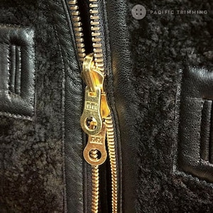 Puede incluir: Primer plano de una cremallera dorada en una chaqueta de cuero negro. La cremallera tiene un tirador de metal con la palabra "Pacific" grabada en él. La chaqueta está hecha de un cuero grueso y duradero y tiene un forro suave y peludo.