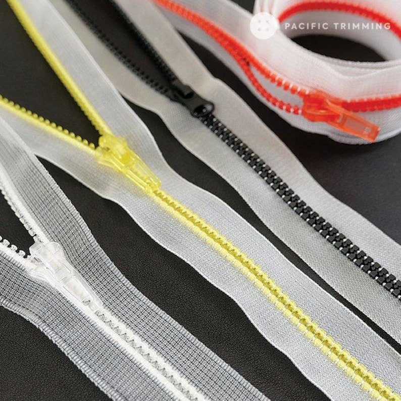 May include: Four different types of zipper tape, including clear, yellow, black, and red. The tape is laid out on a black surface. The text "PACIFIC TRIMMING" is visible in the top right corner of the image.