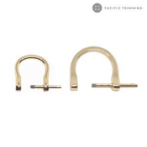 Könnte beinhalten: Zwei goldfarbene D-Ringe aus Metall mit einem Drehriegel. Die D-Ringe sind unterschiedlich groß. Der größere D-Ring hat einen Durchmesser von etwa 5 cm. Der kleinere D-Ring hat einen Durchmesser von etwa 2,5 cm. PACIFIC TRIMMING ist in der oberen rechten Ecke des Bildes gedruckt.