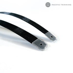 May include: Close-up of two black woven straps with clear plastic end caps. The straps are slightly curved and angled towards the viewer. The text "PACIFIC TRIMMING" is in the upper right corner.