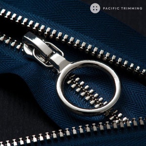 Op de afbeelding: Close-up van een zilveren rits-schuif met een ring, bevestigd aan een blauwe ritsband. De ritsband heeft een visgraatpatroon. De tekst "PACIFIC TRIMMING" is zichtbaar in de rechterbovenhoek van de afbeelding.