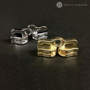 Puede incluir: Dos cierres metálicos, uno plateado y otro dorado, con un diseño ondulado. Los cierres son de metal y tienen un acabado brillante.