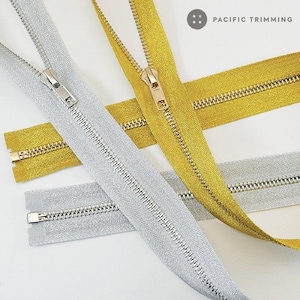 May include: Two zipper tapes, one gold and one silver, with metal teeth and pulls. The zipper tapes are made of a textured fabric with a subtle sheen. The image includes the text "PACIFIC TRIMMING".