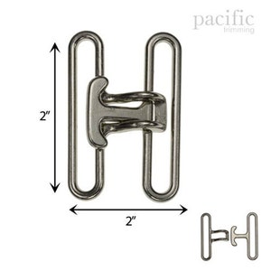 Könnte beinhalten: Silberne Metallschnalle mit zwei Schlaufen, 5 cm lang und breit. Die Schnalle hat ein einzigartiges Design mit einem Mittelstück, das eine einfache Befestigung ermöglicht.