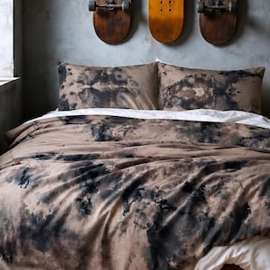 May include: A bedding set featuring a duvet cover and two pillowcases with a dark brown and black tie-dye pattern. The fabric displays a mottled, cloud-like aesthetic. A white flat sheet is visible underneath the duvet, which is draped over a bed.