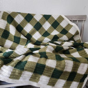 May include: A handmade crochet blanket with a checkered pattern in white, olive green, and dark green. The blanket is draped over a wooden chair, showcasing its texture and design. The blanket's border is a solid white.