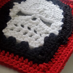 May include: A crocheted square with a white skull design in the center. The skull is surrounded by a black border and a red border.