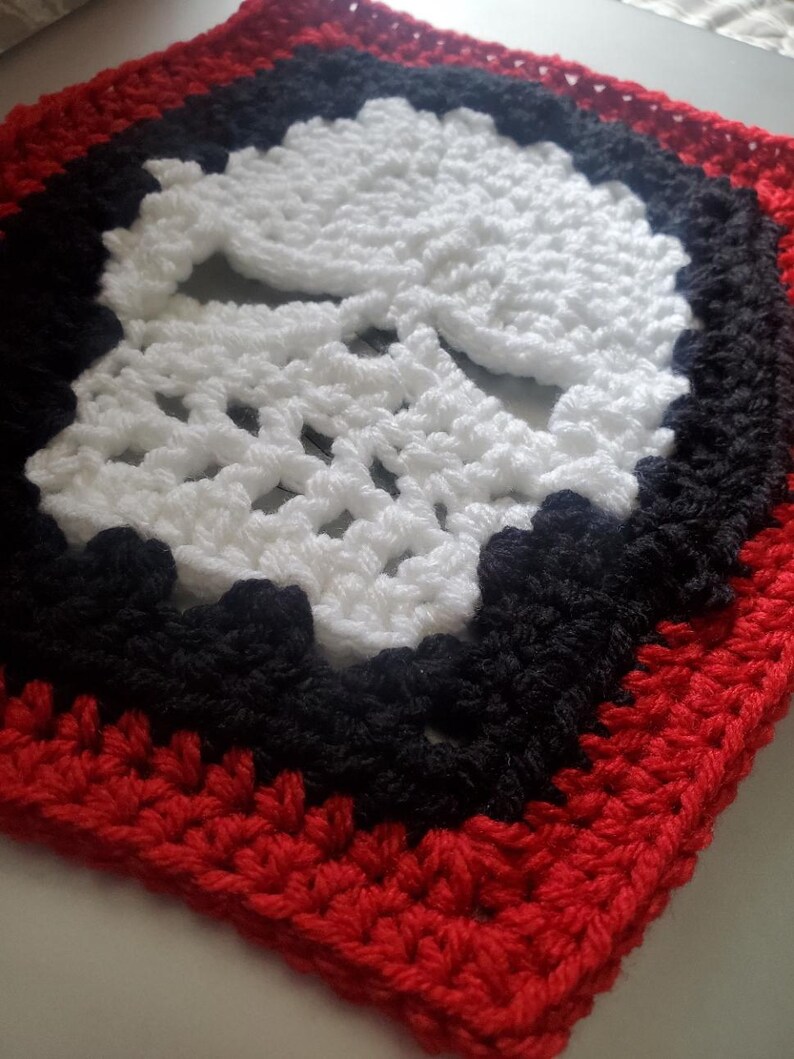 May include: A crocheted square with a white skull design in the center. The skull is surrounded by a black border and a red border.