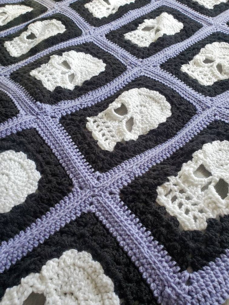 May include: A crocheted blanket with a repeating pattern of white skull motifs on a black and purple background.